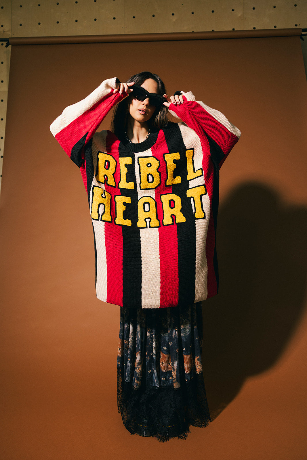 Woman wearing an oversized striped sweater with 'Rebel Heart' text in patches on the front on a brown background.