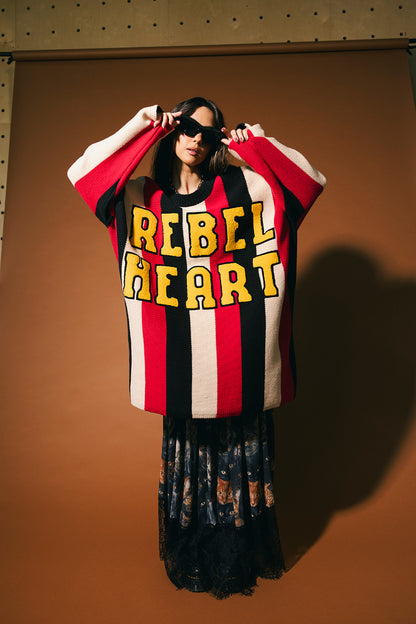 Woman wearing an oversized striped sweater with 'Rebel Heart' text in patches on the front on a brown background.