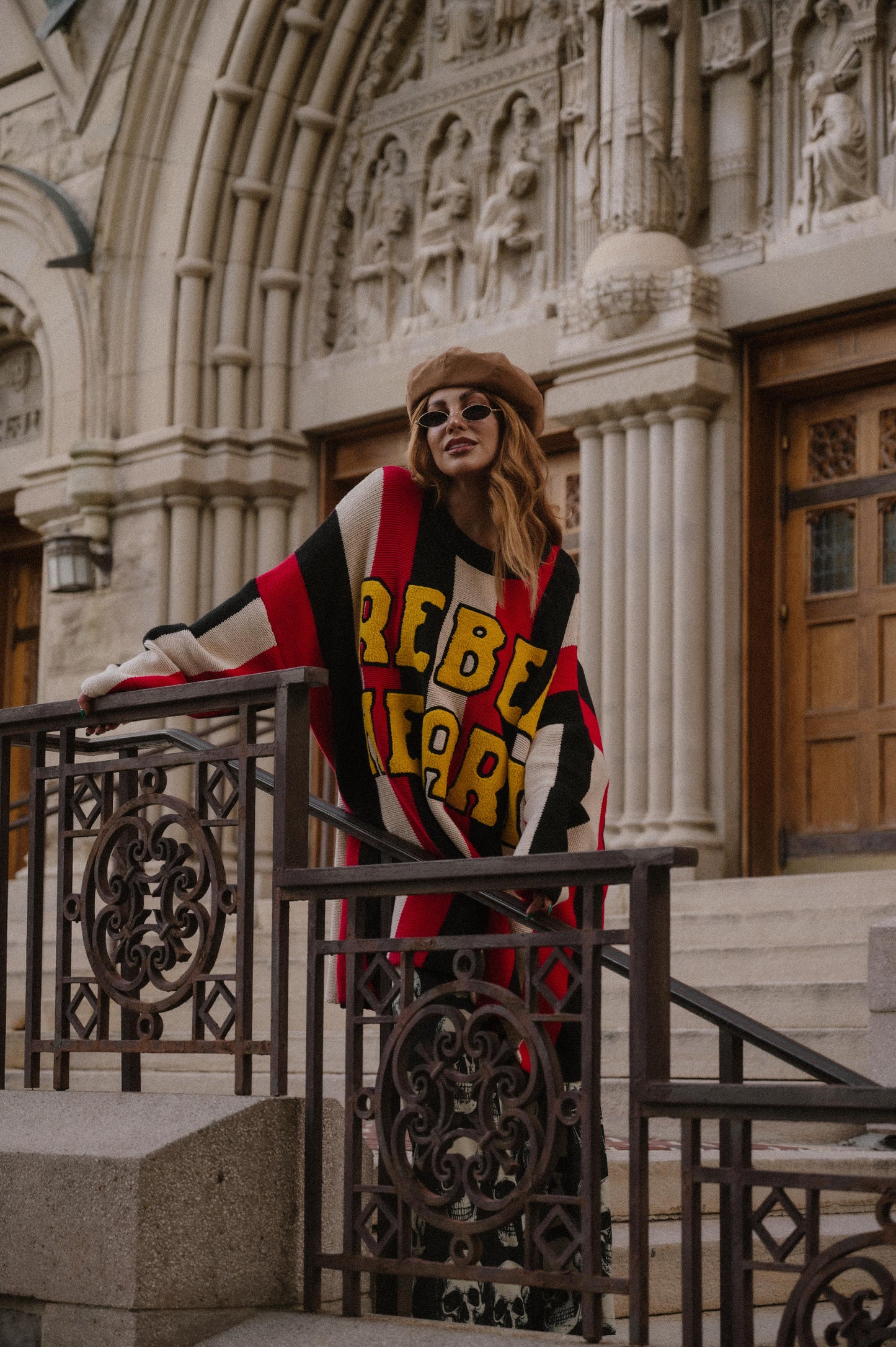 Woman wearing an oversized striped sweater with 'Rebel Heart' text in patches in front of a decorative building entrance.