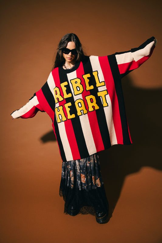 Woman wearing an oversized striped sweater with 'Rebel Heart' in patches on the front on a brown background.