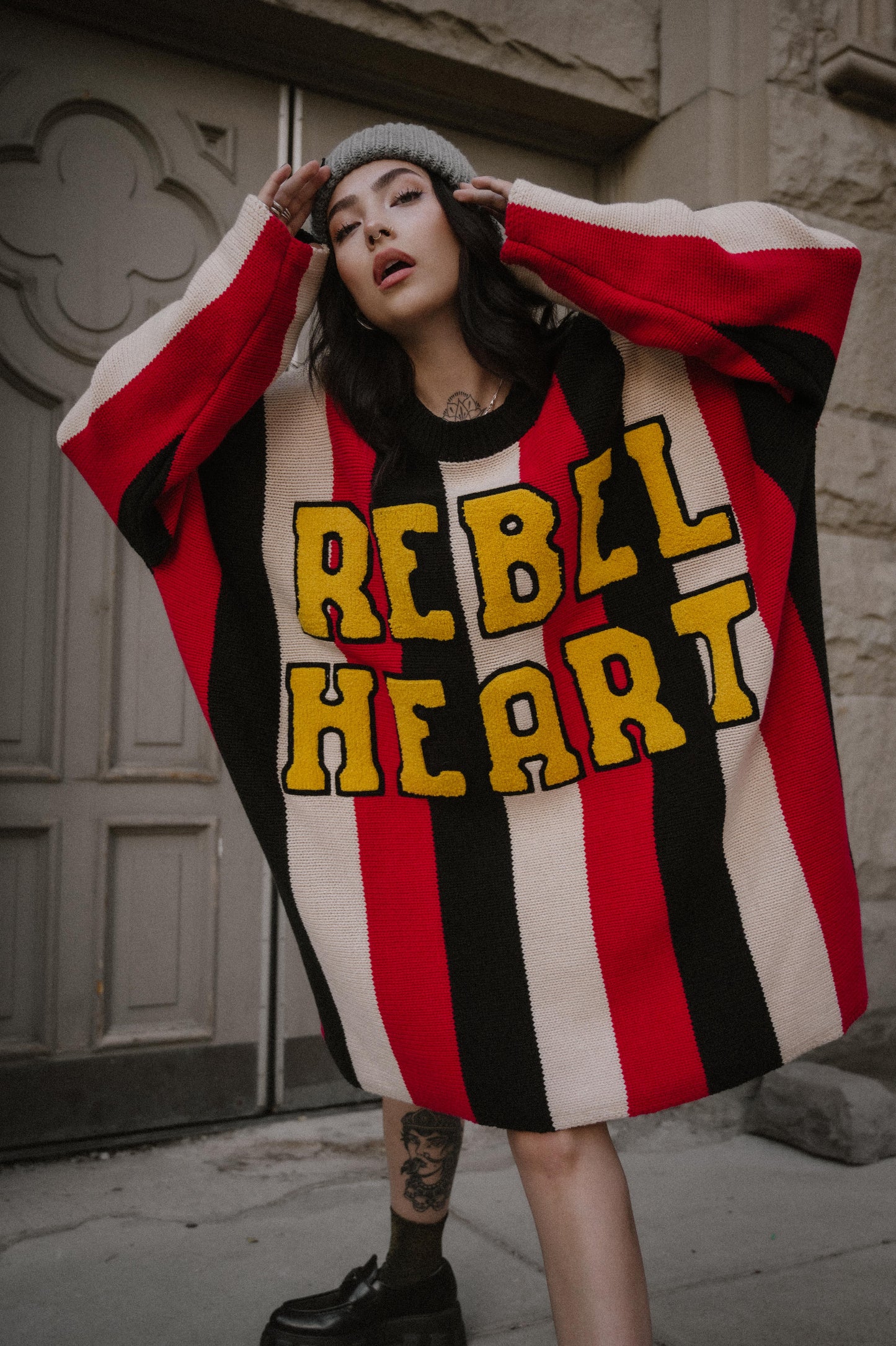 Person wearing an oversized striped sweater with 'REBEL HEART' text in front of a building.