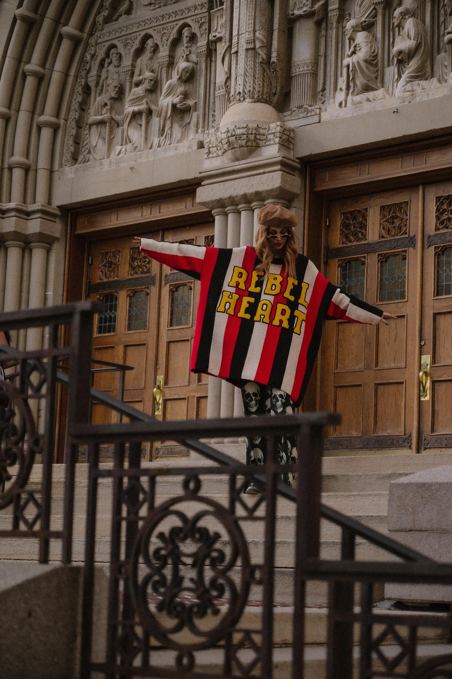 Person wearing an oversized striped sweater with 'REBEL HEART' text in front of a large building entrance.