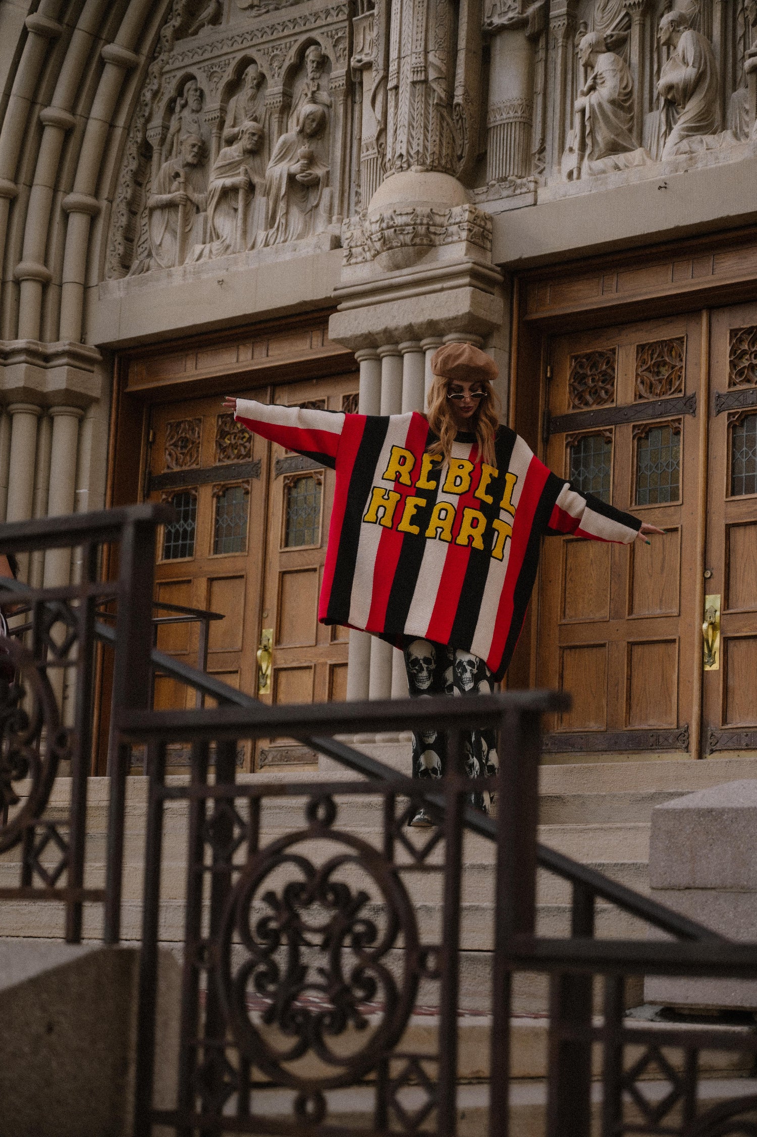 Person wearing an oversized striped sweater with 'REBEL HEART' text in front of a large building entrance.