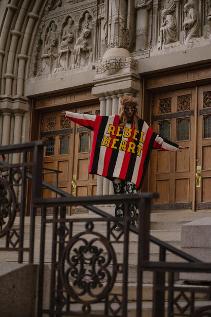 Person wearing an oversized striped sweater with 'REBEL HEART' text in front of a large building entrance.