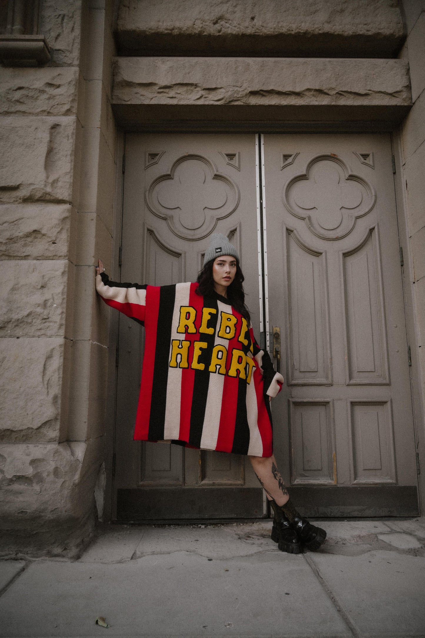 Person wearing an oversized striped sweater with 'REBEL HEART' text in patches in front of a stone building with large doors.