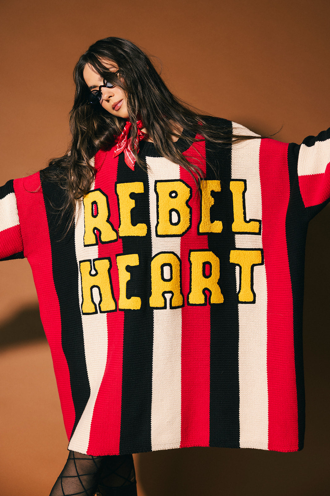 Person wearing an oversized red, black, and white striped sweater with 'REBEL HEART' text in patches on a brown background