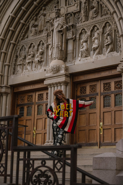 Person wearing an oversized striped sweater with 'Rebel Heart' text in patches standing in front of ornate wooden doors.