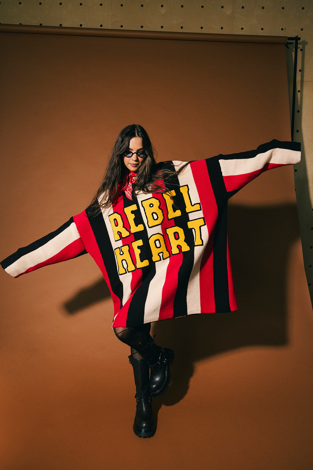 Person wearing an oversized striped sweater with 'REBEL HEART' text in patches against a brown background