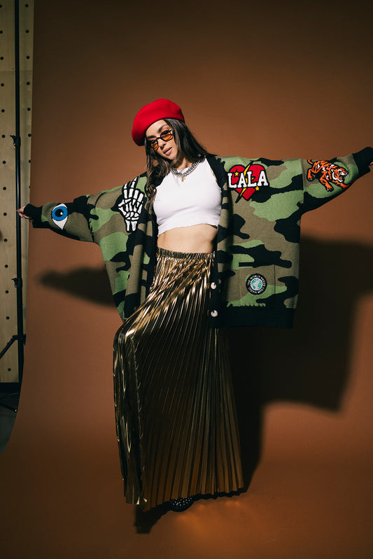 Person wearing a camouflage cardigan with patches, white top, and gold skirt on a brown background