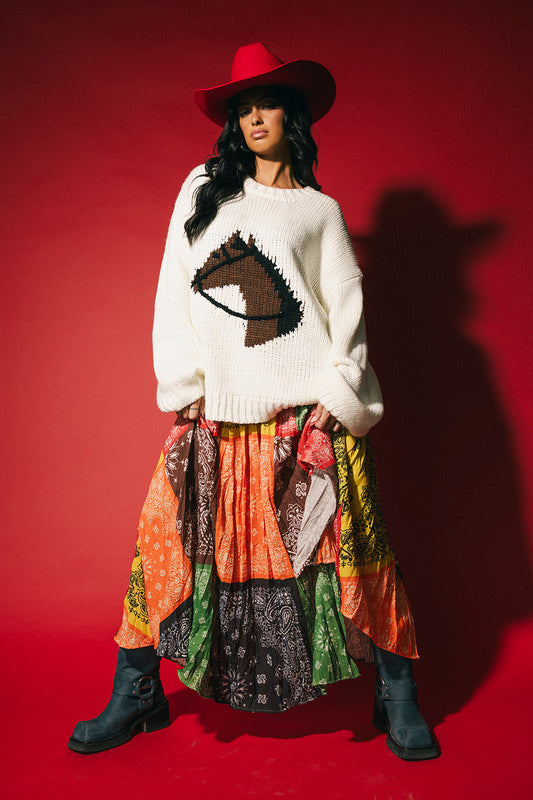 Woman wearing a white sweater with a horse design, colorful skirt, and red cowboy hat on a red background
