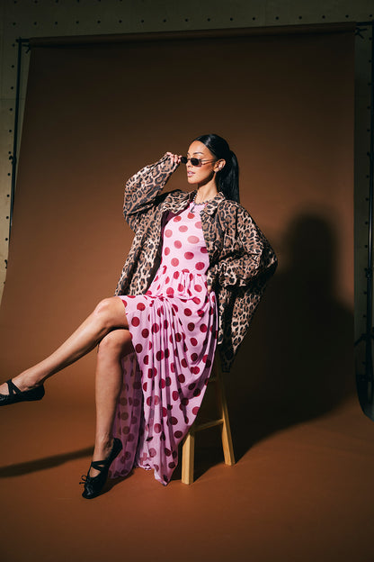 Woman in a polka dot dress and leopard print button up sitting on a stool against a brown background