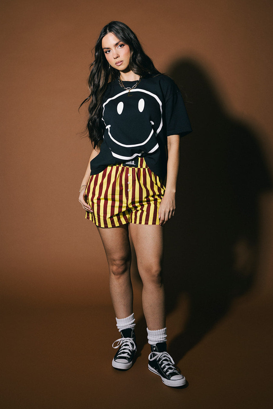 Person wearing a black t-shirt with a smiley face design, yellow and red striped shorts, and black sneakers on a brown background.