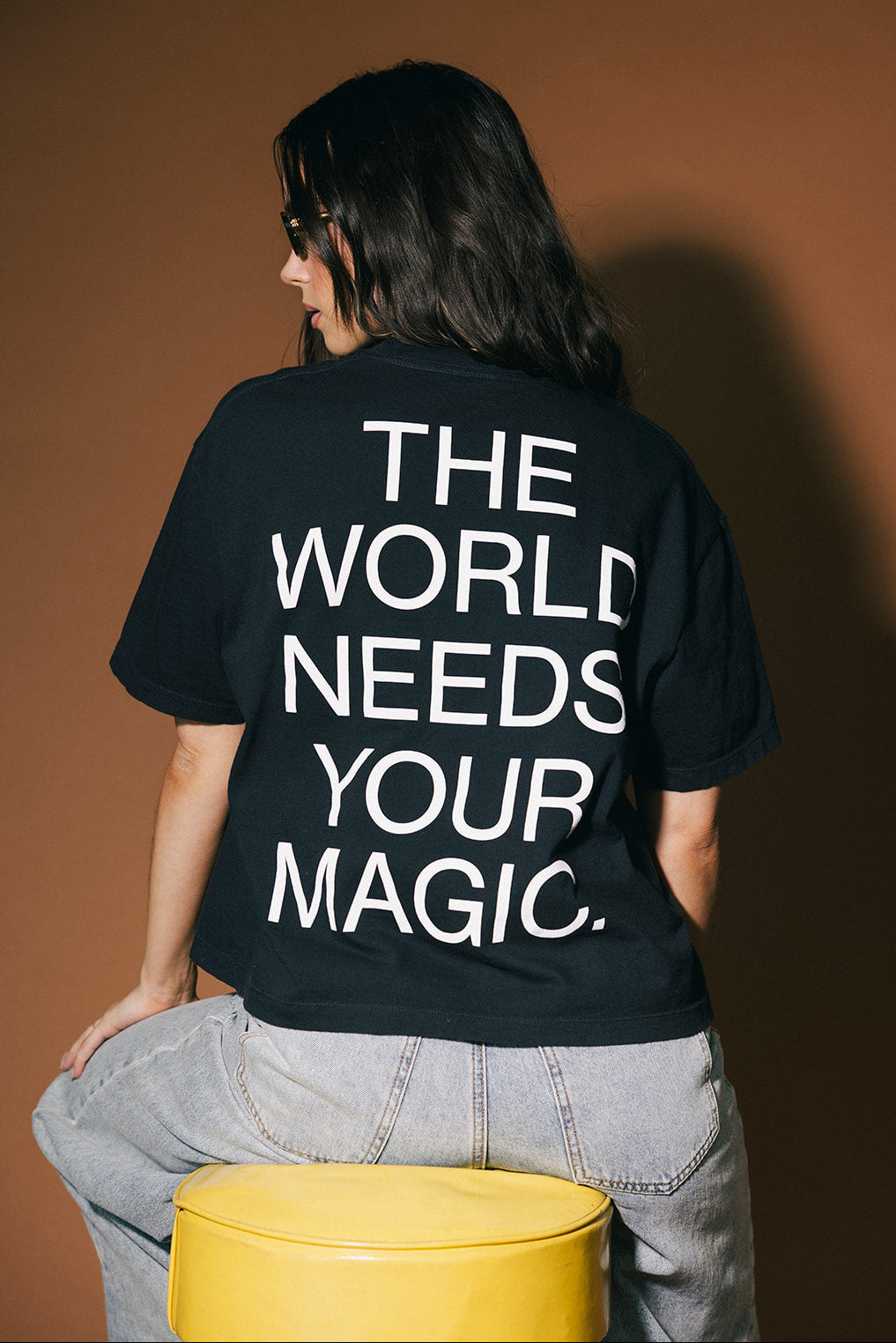 Person wearing a black t-shirt with 'The World Needs Your Magic' text on the back, sitting on a yellow stool against a brown background.