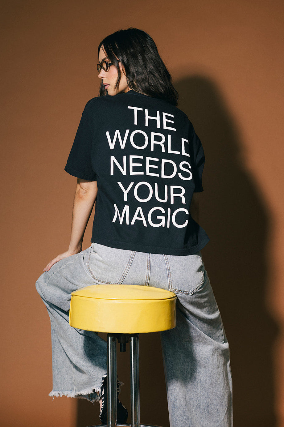 Person wearing a black t-shirt with 'The World Needs Your Magic' text, standing against a brown background.