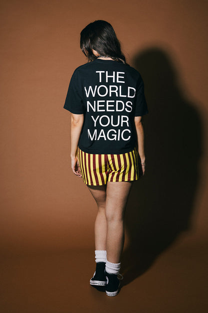Person wearing a black t-shirt with 'The World Needs Your Magic' text on a brown background