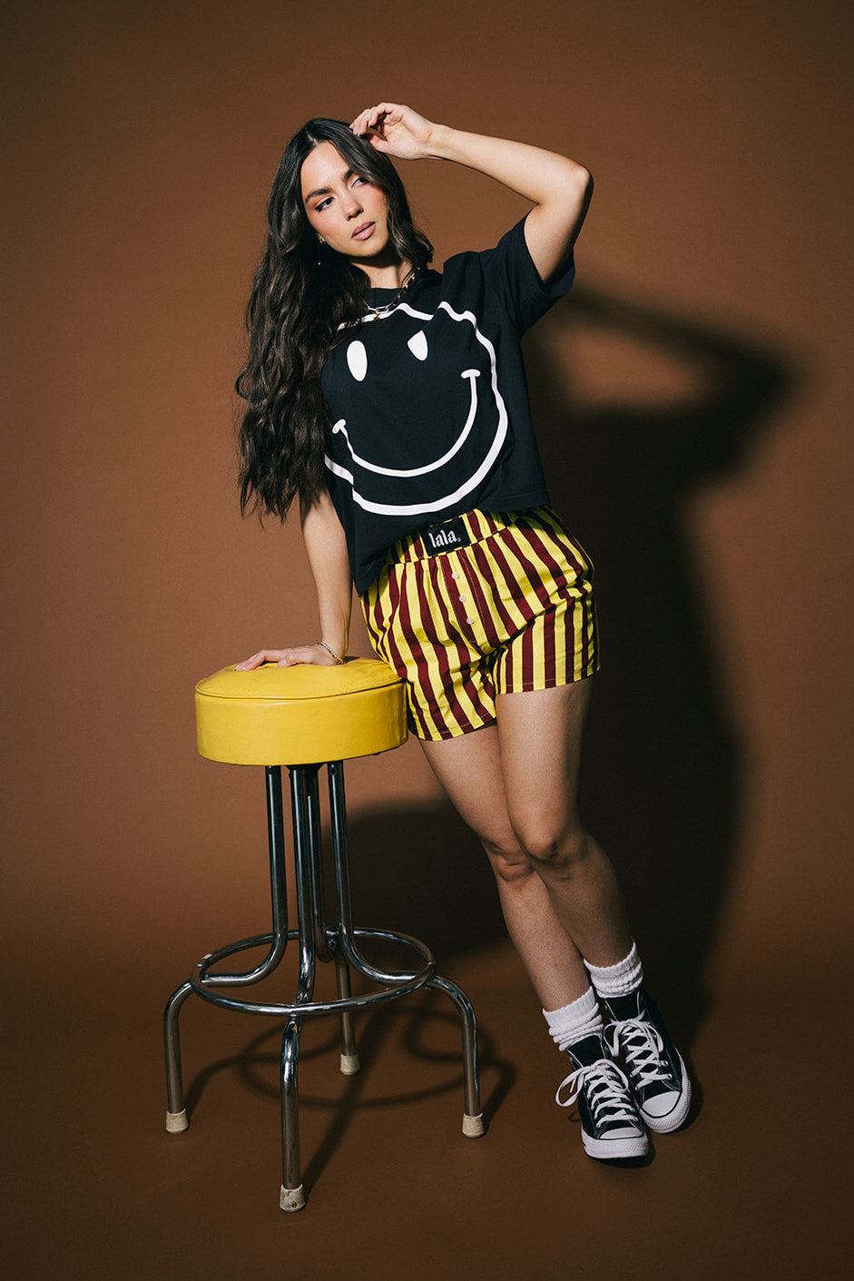 Person wearing a black smiley face shirt and yellow and red striped shorts standing next to a yellow stool on a brown background.