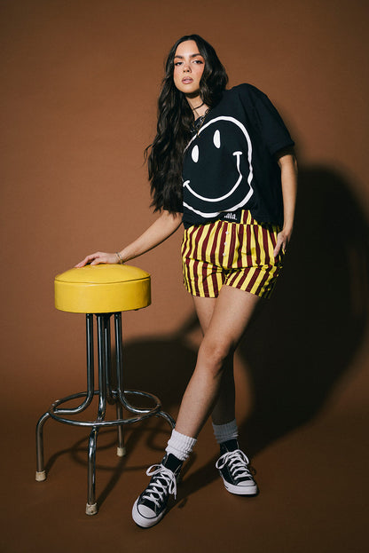 Woman in black smiley face t-shirt and yellow striped shorts standing next to a yellow stool on a brown background
