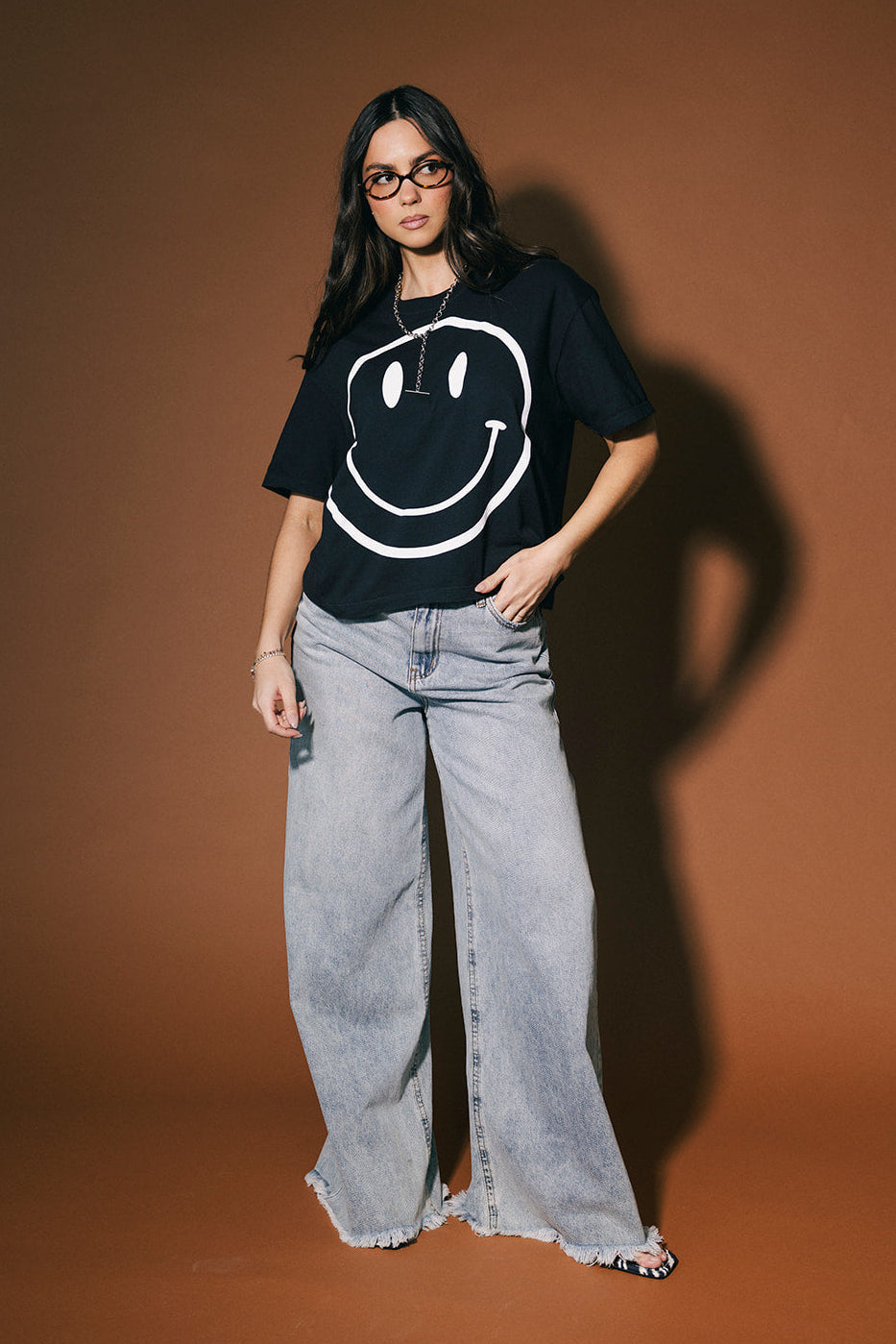 Person wearing a black t-shirt with a smiley face design and light blue jeans against a brown background