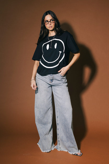 Person wearing a black t-shirt with a smiley face design and light blue jeans against a brown background
