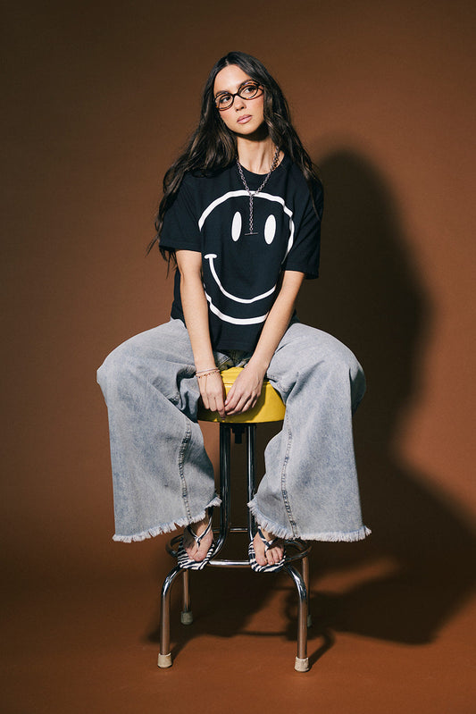Person sitting on a stool wearing a black t-shirt with a smiley face design against a brown background