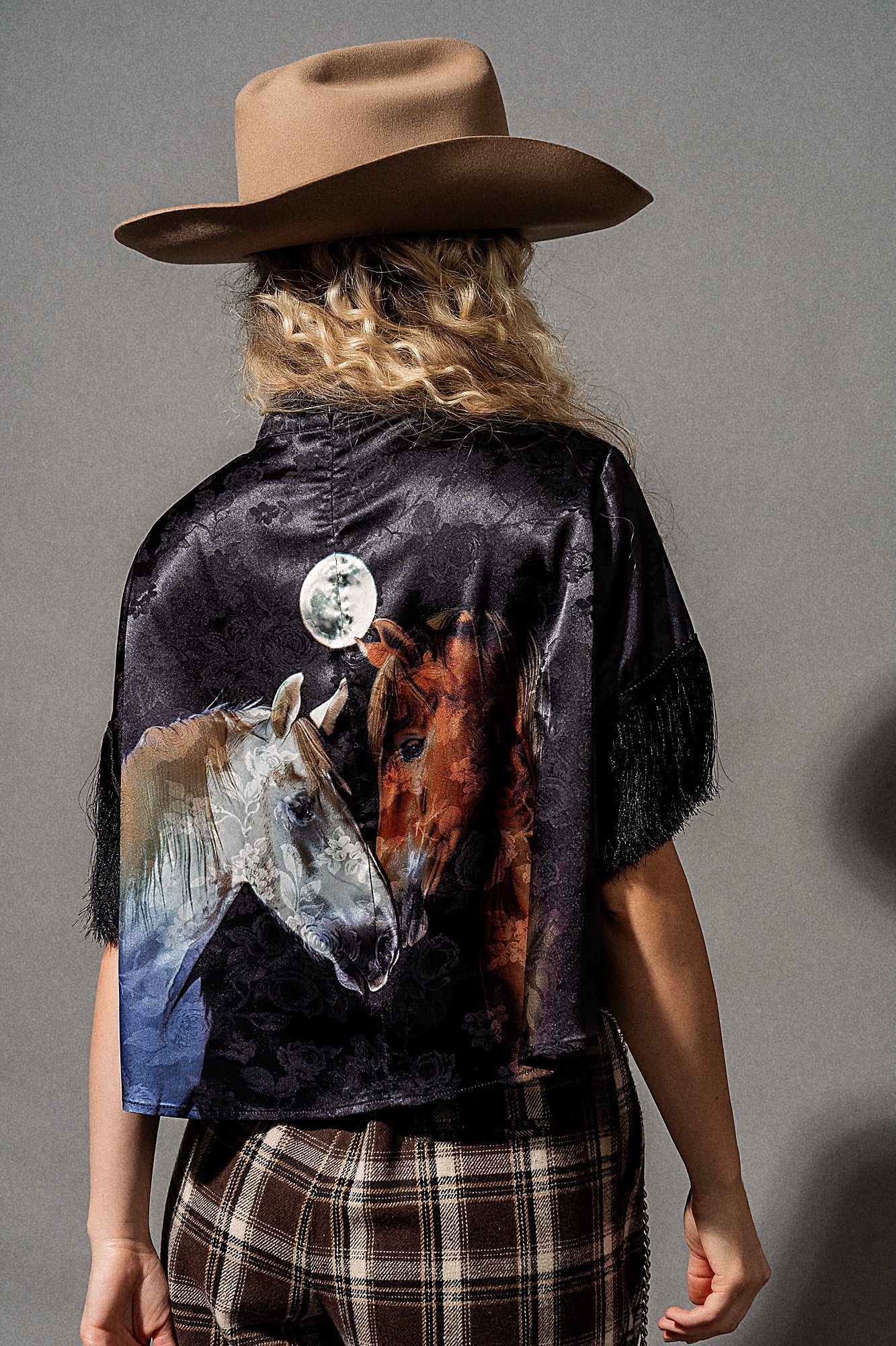 Person wearing a black shirt with a horse design, plaid pants, and a cowboy hat on a gray background