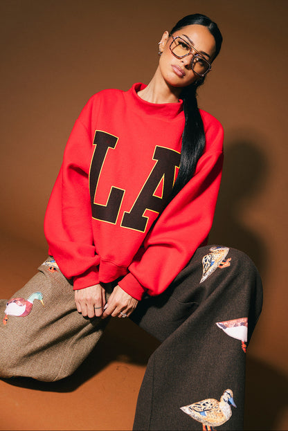 Person wearing a red sweatshirt with 'LA' and half and half pants with patches on a brown background