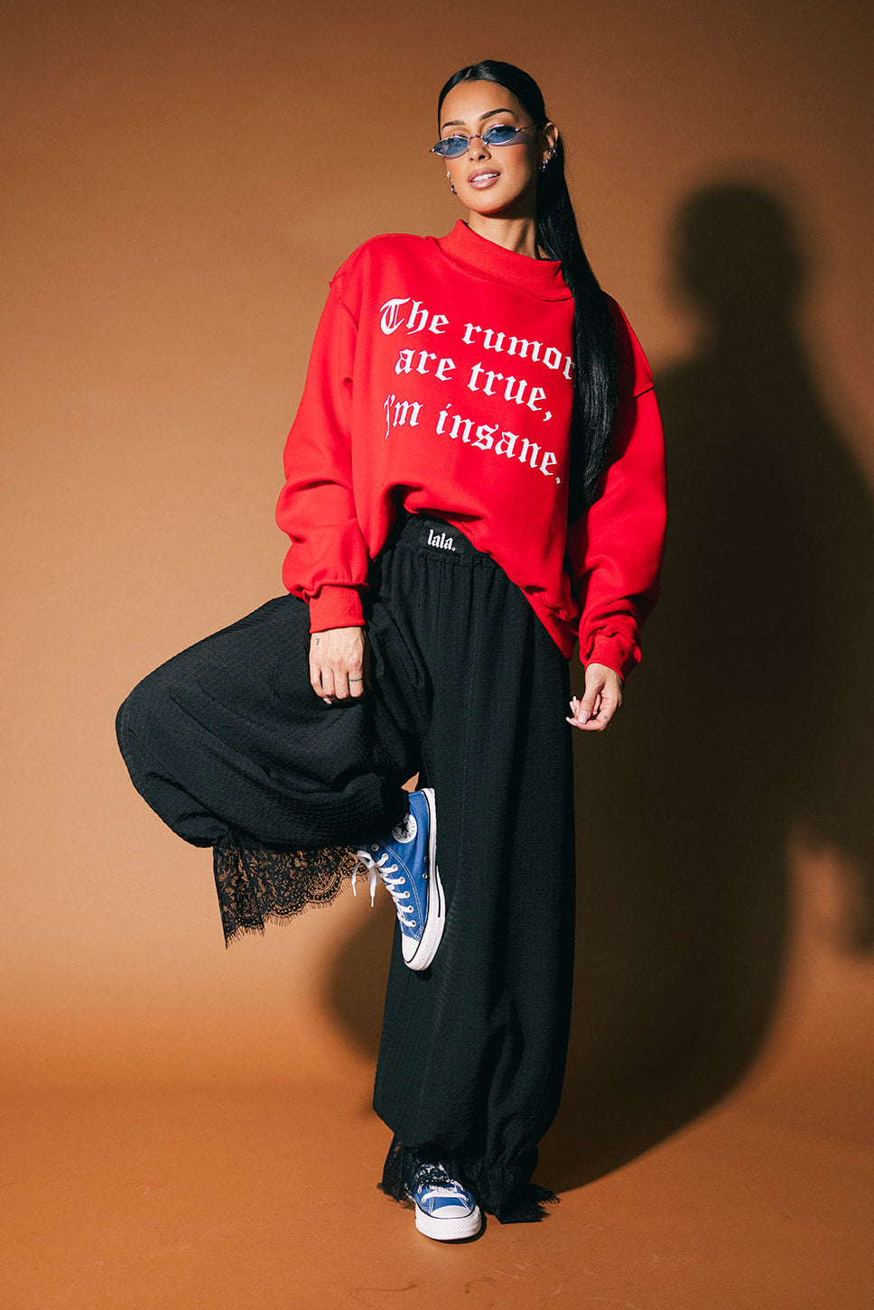 Person wearing a red sweatshirt with text, black pants, and blue sneakers on a brown background