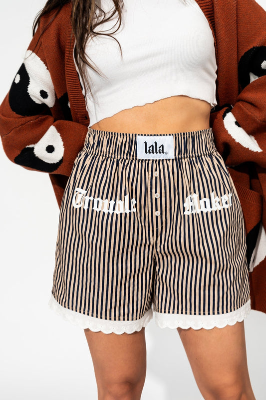 photo of girl wearing tan and navy stripe boxer shorts that say "trouble maker" across the front in white and has white lace trim on the hem with bows, elastic waistband, side pockets, 3 button faux fly, and white label that says "lala" on the waistband