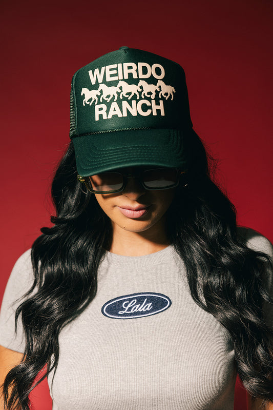 Person wearing a green trucker hat with 'Weirdo Ranch' text and horses in puff print and sunglasses with a gray shirt featuring a logo.