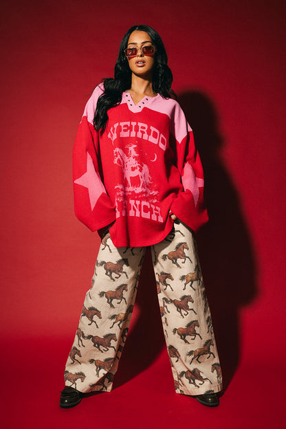 Person wearing a red and pink waffle knit hockey jersey inspired top with 'Weirdo Ranch' text and cowgirl riding a unicorn graphic on the front and star graphics on the sleeves on a red background