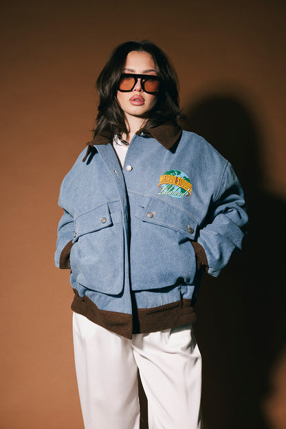 Person wearing a blue denim jacket with a logo, white pants, and sunglasses against a brown background