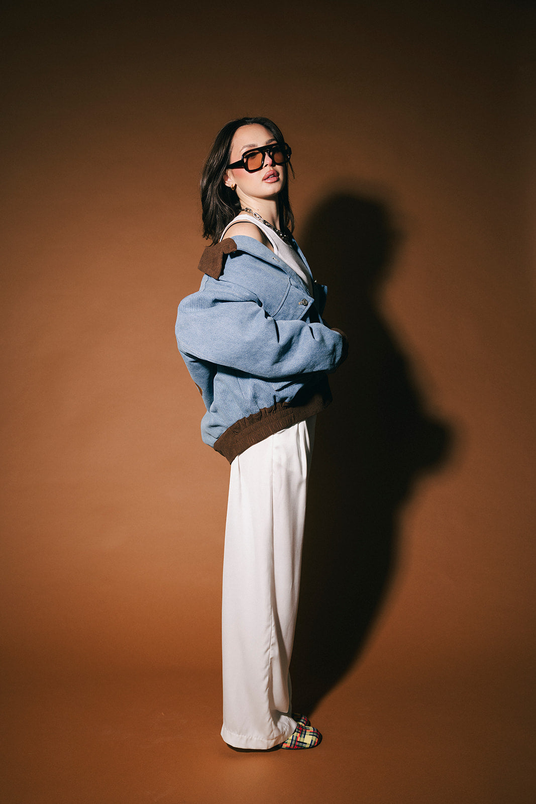Person wearing a blue denim jacket and sunglasses against a brown background