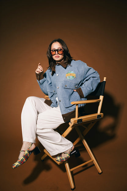 Person sitting on a director's chair wearing a blue denim jacket and sunglasses against a brown background