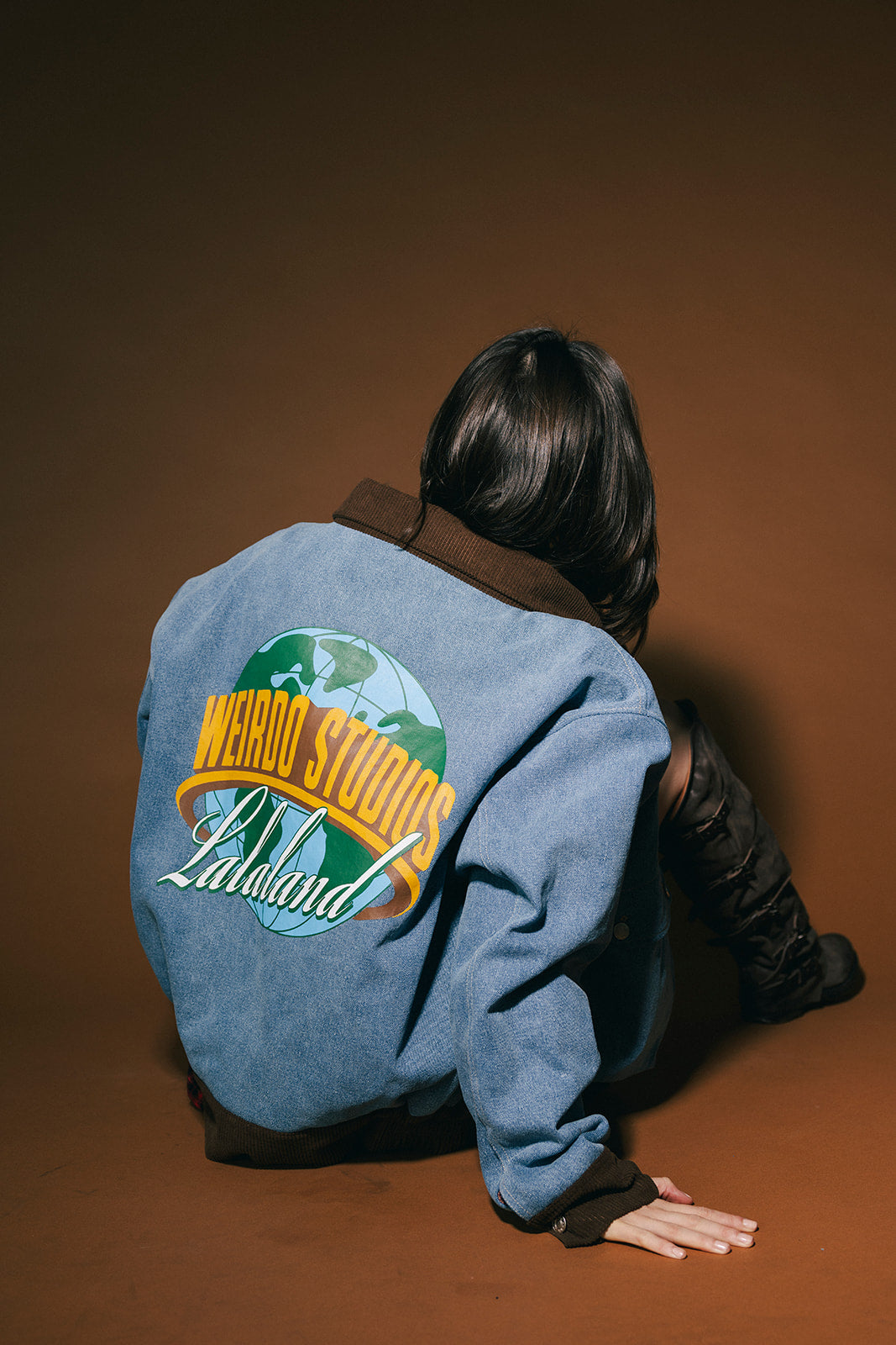 Person wearing a blue denim jacket with 'Weirdo Studios' logo on the back against a brown background