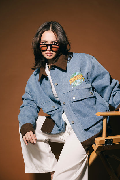 Person wearing a blue denim jacket with a logo, white pants, and sunglasses on a brown background