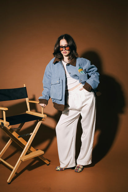 Person wearing a blue denim jacket and white pants standing next to a director's chair against a brown background