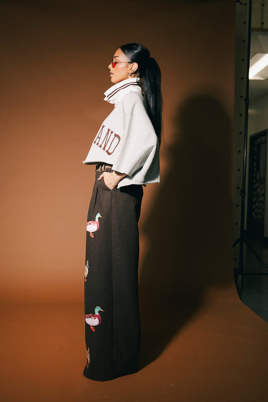 Person wearing a grey cropped polo with text and dark pants with duck patches on a brown background