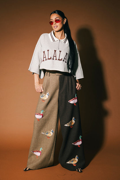Woman wearing a grey cropped polo with 'LALALAND' text and brown wide-leg pants with duck patches on a brown background.