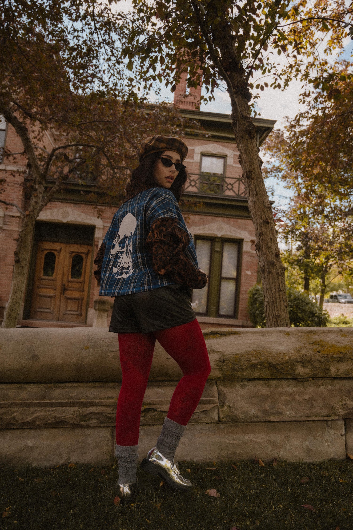 Person wearing a blue plaid top and skull on the back with leopard print sleeves and collar and red tights standing in front of an old building.