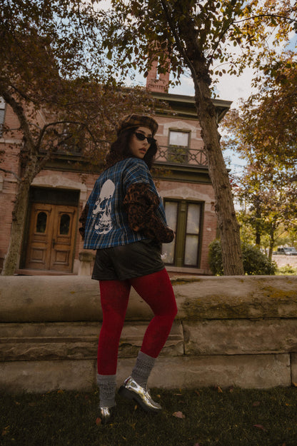 Person wearing a blue plaid top and skull on the back with leopard print sleeves and collar and red tights standing in front of an old building.