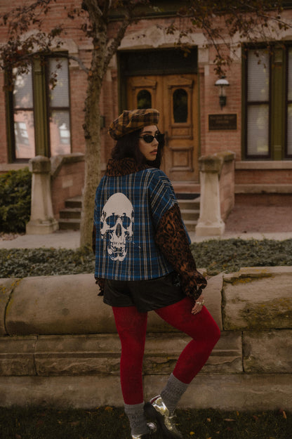 Person wearing a blue plaid shirt with a skull design and leopard sleeves and collars, red tights, and sunglasses standing in front of a brick building.