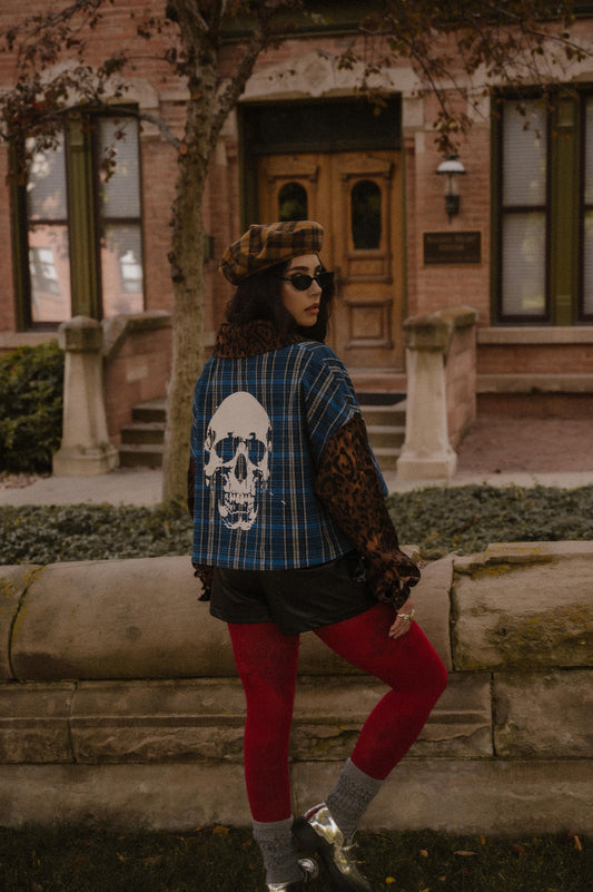 Person wearing a blue plaid shirt with a skull design and leopard sleeves and collars, red tights, and sunglasses standing in front of a brick building.