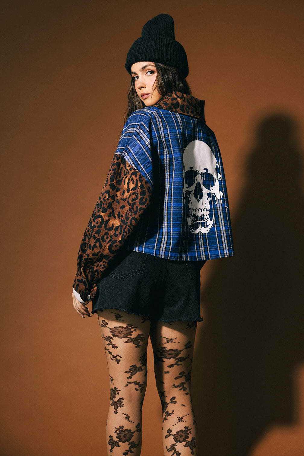 Person wearing a blue plaid shirt with leopard print sleeves and a skull design, standing against a brown background.