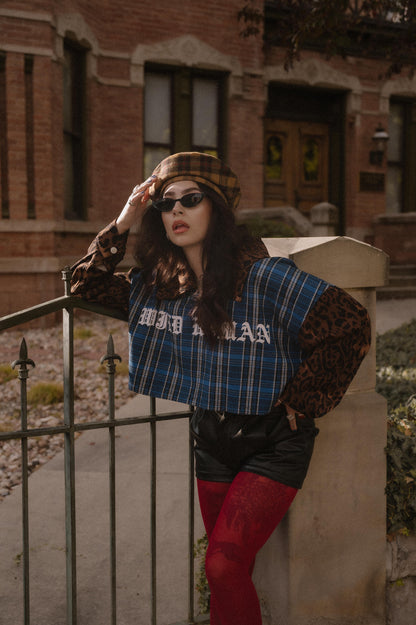 Person wearing a blue plaid shirt with 'Wild Woman' text on the front and leopard sleeves and collar, sunglasses, and red tights standing in front of a brick building.