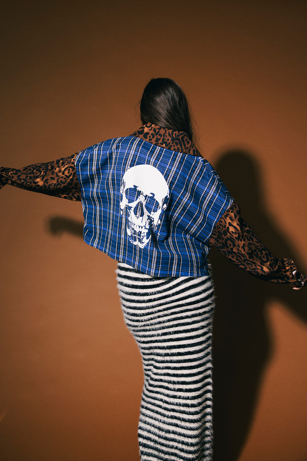 Woman wearing a blue plaid top with a skull on the back and leopard print sleeves and collar against a brown background.