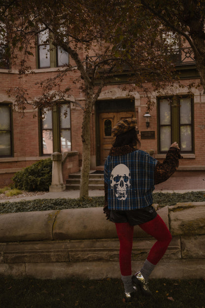 Person wearing a blue plaid shirt with a skull design and leopard print sleeves and collar standing in front of a brick building.