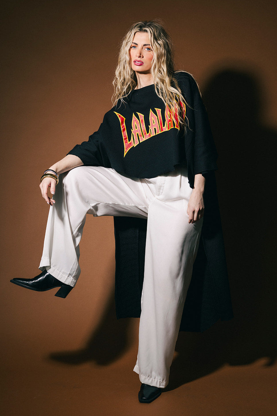 Person wearing a black sweater with 'LALALAND' text, white pants, and black shoes on a brown background