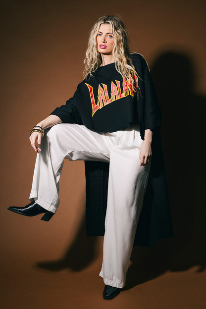 Person wearing a black sweater with 'LALALAND' text, white pants, and black shoes on a brown background