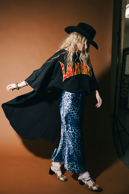 Person wearing a black coat with 'Leather' text, blue leopard print skirt, and black hat against a brown background