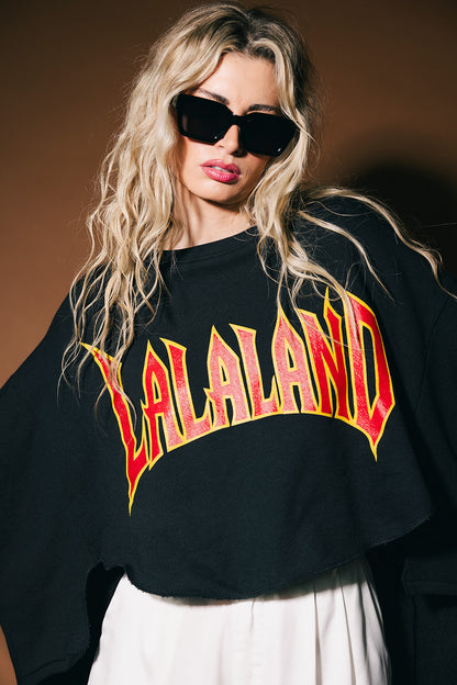 Person wearing a black sweatshirt with 'LALALAND' text, sunglasses, and white pants against a brown background